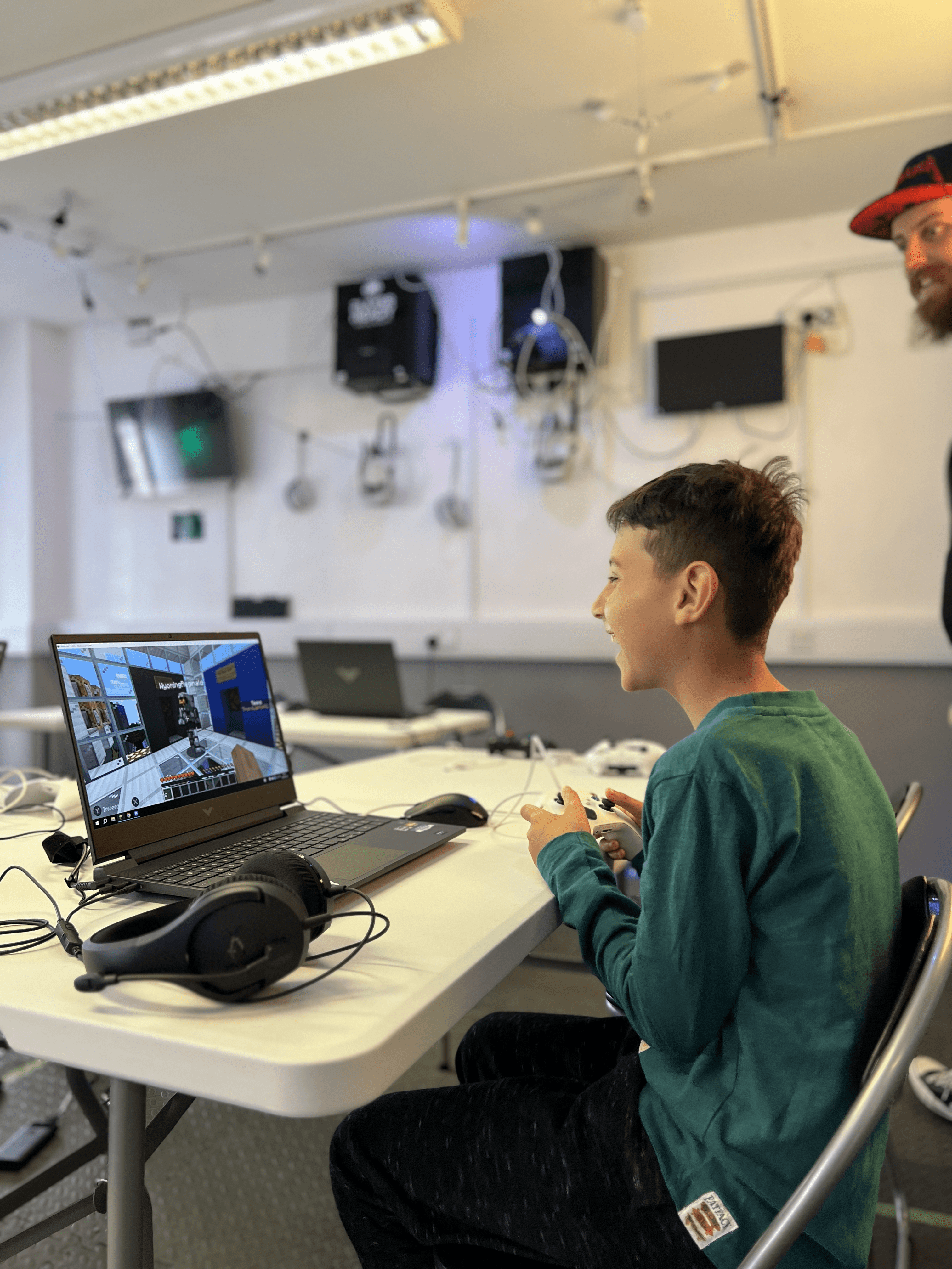 A young person smiling and engaged during a mentoring session at Player Ready, working on a laptop with VR headsets visible in the background